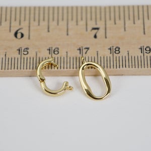 May include: Two gold-colored jewelry clasps. One is a curved, hinged clasp, and the other is an oval-shaped pendant. The clasps are on a white surface next to a wooden ruler with inch markings.