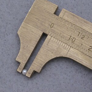 May include: A close-up of a white pearl bead measured with a brass caliper. The caliper reads 10 millimeters.