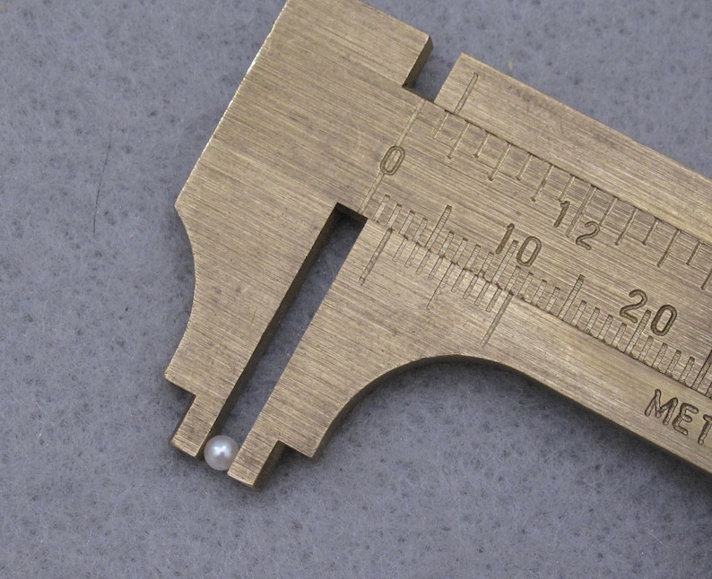 May include: A close-up of a white pearl bead measured with a brass caliper. The caliper reads 10 millimeters.
