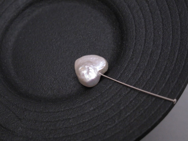 May include: A white heart-shaped pearl on a black plate with a silver pin attached.