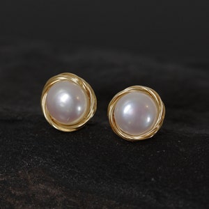 Può includere: Un paio di orecchini a perno con perle. Ogni orecchino presenta una perla rotonda e lucente incastonata in una montatura in metallo dorato ritorto. Gli orecchini sono esposti su uno sfondo scuro e strutturato, evidenziando il loro design elegante.