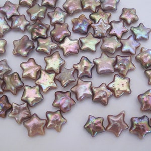 May include: A collection of iridescent, star-shaped pearl beads in shades of lavender and pink. The beads are scattered on a white surface.