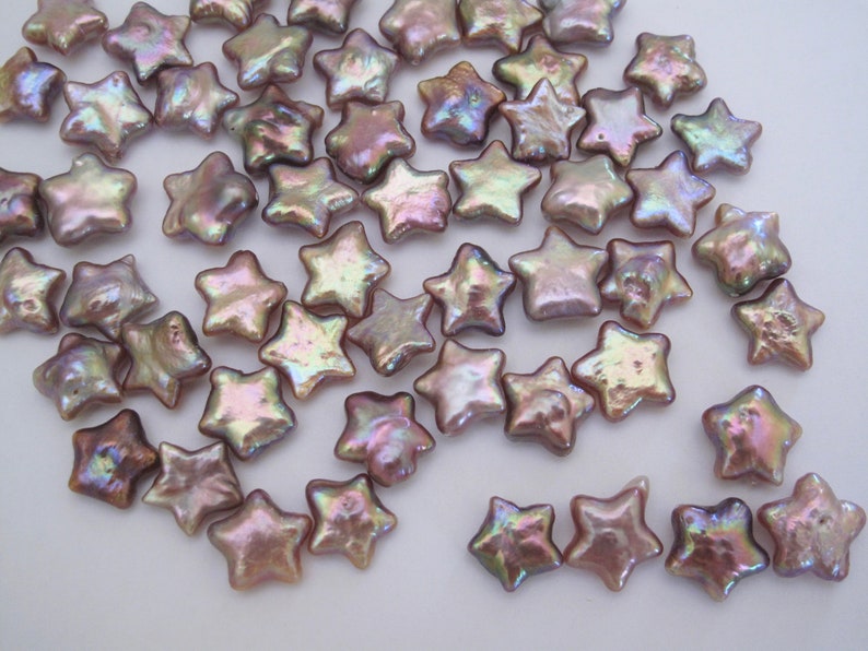 May include: A collection of iridescent, star-shaped pearl beads in shades of lavender and pink. The beads are scattered on a white surface.