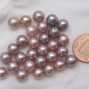 May include: A group of approximately 25 iridescent purple and pink freshwater pearls. The pearls are round and smooth, and they are arranged in a loose pile. A US penny is included for scale.