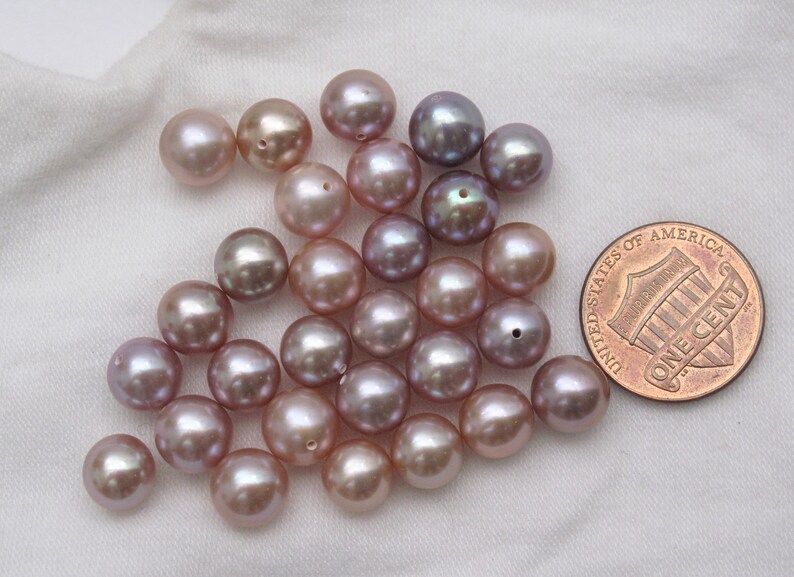 May include: A group of approximately 25 iridescent purple and pink freshwater pearls. The pearls are round and smooth, and they are arranged in a loose pile. A US penny is included for scale.