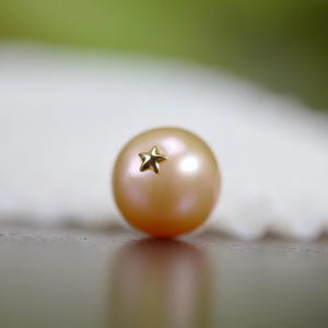May include: A close-up of a peach-colored pearl with a small gold star affixed to its surface. The pearl is round and has a subtle sheen. The background is blurred, with hints of white and green.