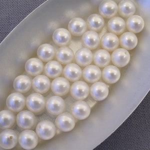 May include: A collection of white round pearls in a white oval dish. The pearls are smooth and shiny.