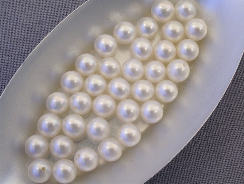 May include: A collection of white round pearls in a white oval dish. The pearls are smooth and shiny.