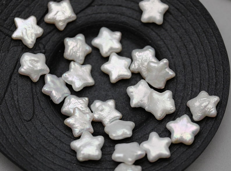 May include: A collection of white pearl star-shaped beads on a black plate. The beads are iridescent and have a smooth, polished finish.