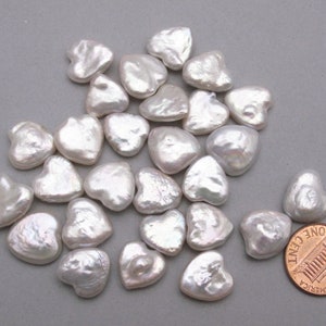May include: A pile of white heart-shaped freshwater pearls, some with a slight iridescent sheen. The pearls are approximately the size of a US penny.