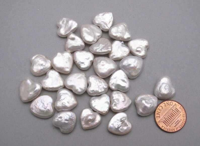 May include: A pile of white heart-shaped freshwater pearls, some with a slight iridescent sheen. The pearls are approximately the size of a US penny.