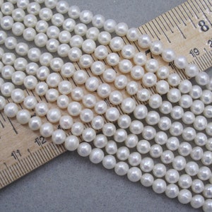May include: A strand of white pearl beads, approximately 6mm in diameter, displayed on a wooden ruler. The ruler shows measurements in inches.