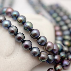 May include: Close-up of multiple strands of dark gray and iridescent pearl necklaces. The pearls have a smooth, round shape and reflect light with hues of green, purple, and blue. The necklaces are displayed on a white surface.