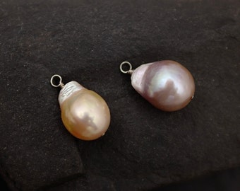 May include: Two baroque pearl pendants with silver-colored findings. One pearl is a creamy peach color, and the other is a mauve color. Both have a pear-like shape and a lustrous surface, set against a dark, textured background.