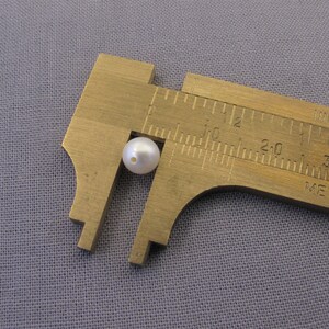 May include: A white pearl bead measured with a metal caliper. The caliper reads 1 inch.