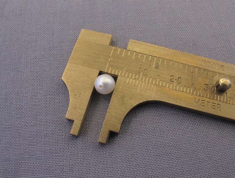May include: A white pearl bead measured with a metal caliper. The caliper reads 1 inch.