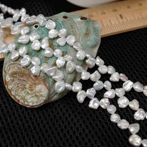 May include: A close-up of a white pearl necklace draped over a teal and brown seashell. The pearls are irregularly shaped and have a lustrous sheen. A wooden ruler is partially visible in the background, set against a black mesh surface.