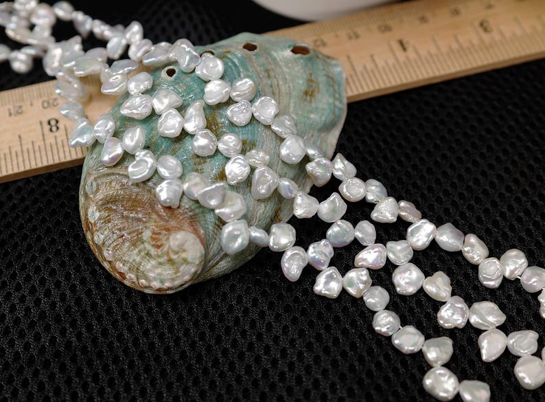 May include: A close-up of a white pearl necklace draped over a teal and brown seashell. The pearls are irregularly shaped and have a lustrous sheen. A wooden ruler is partially visible in the background, set against a black mesh surface.