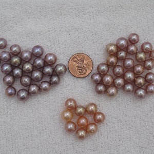 May include: A collection of lustrous, round pearls in shades of pink, peach, and lavender. The pearls are scattered on a white surface, with a penny included for size reference. The pearls vary in color and size.