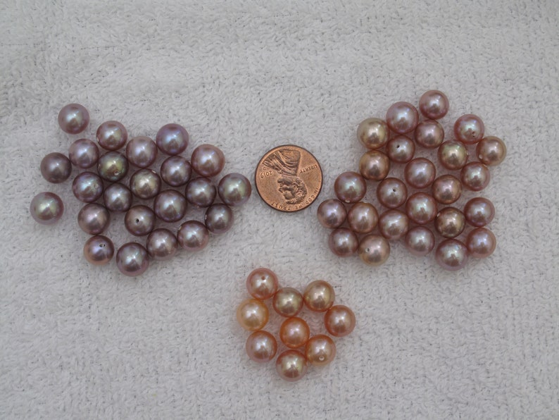 May include: A collection of lustrous, round pearls in shades of pink, peach, and lavender. The pearls are scattered on a white surface, with a penny included for size reference. The pearls vary in color and size.