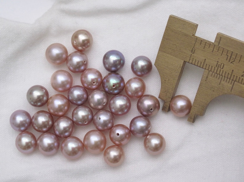 May include: A group of 20 light purple and lavender pearls with a ruler measuring one of the pearls at 10 mm. The pearls are round and have a smooth, glossy finish.