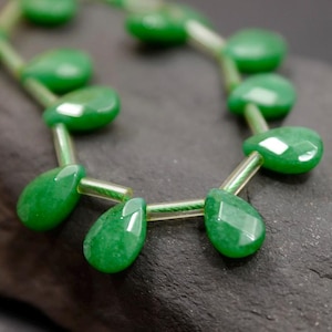 May include: A strand of green gemstone beads in a teardrop shape. The beads are strung on clear iridescent thread.