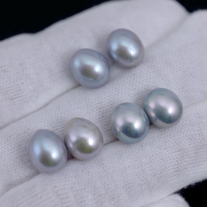 Puede incluir: Seis perlas ovaladas y brillantes en tonos grises y plateados se muestran sobre una tela blanca. Las perlas tienen una superficie lisa e iridiscente, que refleja la luz. Las perlas están dispuestas en un patrón disperso.