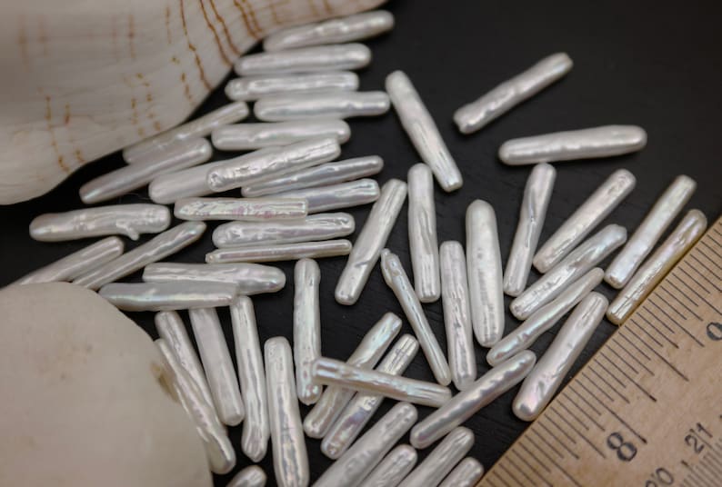 Puede incluir: Una colecci&oacute;n de cuentas blancas e iridiscentes, de forma rectangular alargada. Las cuentas est&aacute;n esparcidas sobre una superficie oscura con una regla en la esquina inferior derecha.