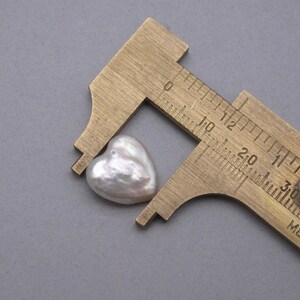 May include: A white heart-shaped pearl measured with a brass caliper. The caliper shows measurements in inches and centimeters.