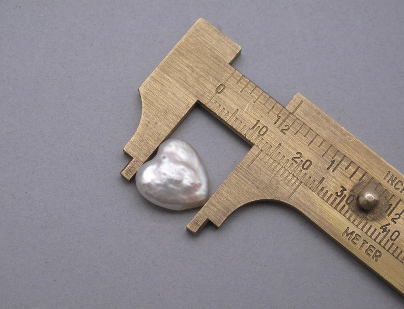 May include: A white heart-shaped pearl measured with a brass caliper. The caliper shows measurements in inches and centimeters.