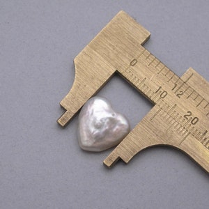 May include: A white heart-shaped pearl measured with a brass caliper. The pearl is approximately 1.2 cm wide.