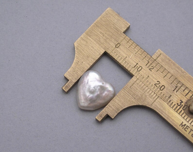 May include: A white heart-shaped pearl measured with a brass caliper. The pearl is approximately 1.2 cm wide.