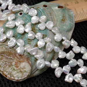May include: A strand of irregular, white freshwater pearls. The pearls are displayed on a multi-colored, iridescent abalone shell. A wooden ruler is visible in the background. The pearls have a lustrous sheen.