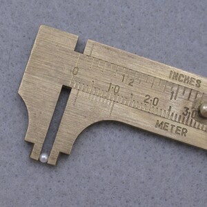 May include: A close-up of a small, white pearl being measured with a brass caliper. The caliper has markings in inches and centimeters.