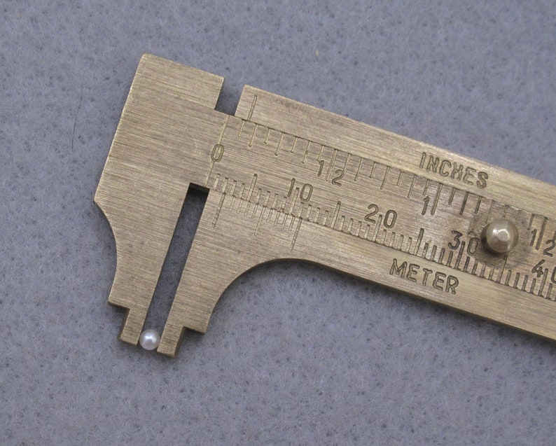 May include: A close-up of a small, white pearl being measured with a brass caliper. The caliper has markings in inches and centimeters.