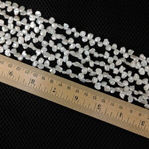 May include: Strands of white, irregular-shaped beads, possibly pearls, are arranged across a black surface. A wooden ruler, marked in inches, is placed diagonally across the beads, providing a size reference.