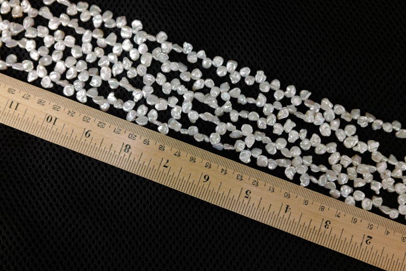 May include: Strands of white, irregular-shaped beads, possibly pearls, are arranged across a black surface. A wooden ruler, marked in inches, is placed diagonally across the beads, providing a size reference.
