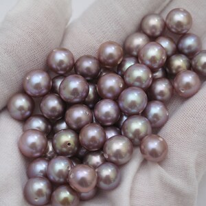 May include: A collection of lustrous, round pearls in varying shades of lavender and gray. The pearls are displayed on a white fabric background, showcasing their smooth, iridescent surfaces. These pearls are ideal for jewelry making.