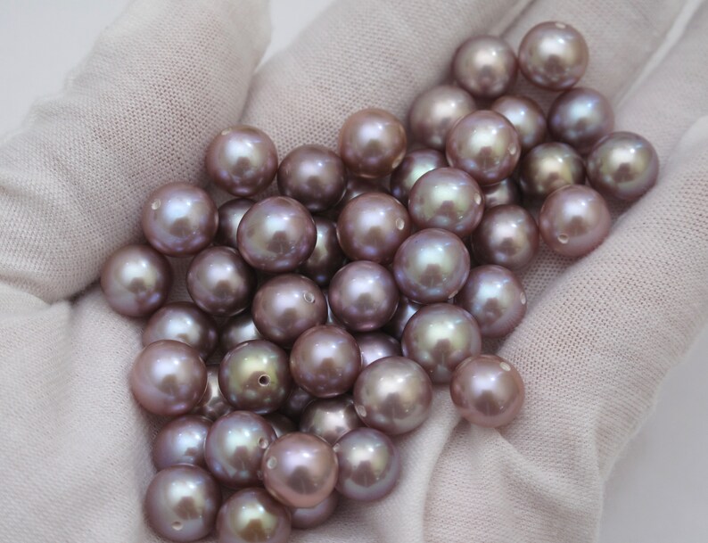 May include: A collection of lustrous, round pearls in varying shades of lavender and gray. The pearls are displayed on a white fabric background, showcasing their smooth, iridescent surfaces. These pearls are ideal for jewelry making.