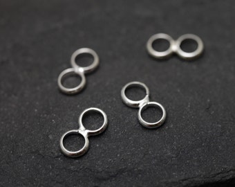 May include: Four small, silver-colored infinity charm connectors are scattered on a dark, textured surface. Each charm features two connected circles, creating an infinity symbol shape. The charms are made of a shiny metal.