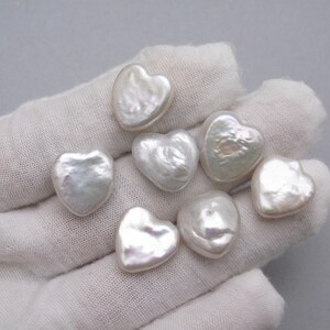 May include: Seven white heart-shaped pearls on a white surface. The pearls are smooth and have a slightly iridescent sheen.