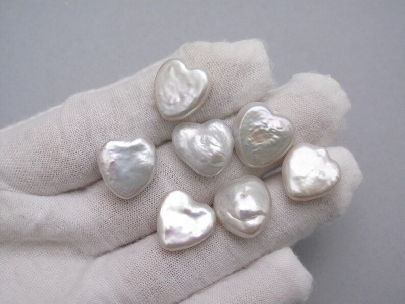 May include: Seven white heart-shaped pearls on a white surface. The pearls are smooth and have a slightly iridescent sheen.