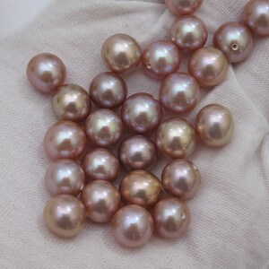 May include: A collection of lustrous, round pearls in varying shades of pink and champagne. The pearls have a smooth, reflective surface, with some showing a slight iridescent sheen. The pearls are scattered on a white fabric background.