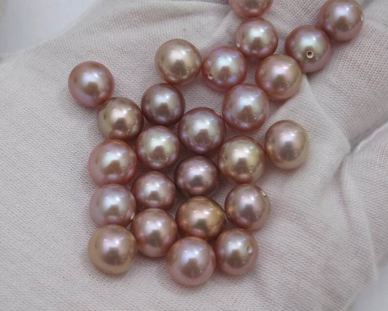 May include: A collection of lustrous, round pearls in varying shades of pink and champagne. The pearls have a smooth, reflective surface, with some showing a slight iridescent sheen. The pearls are scattered on a white fabric background.