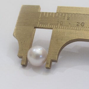 May include: A white pearl is measured with a brass caliper. The pearl is approximately 10 millimeters in diameter.
