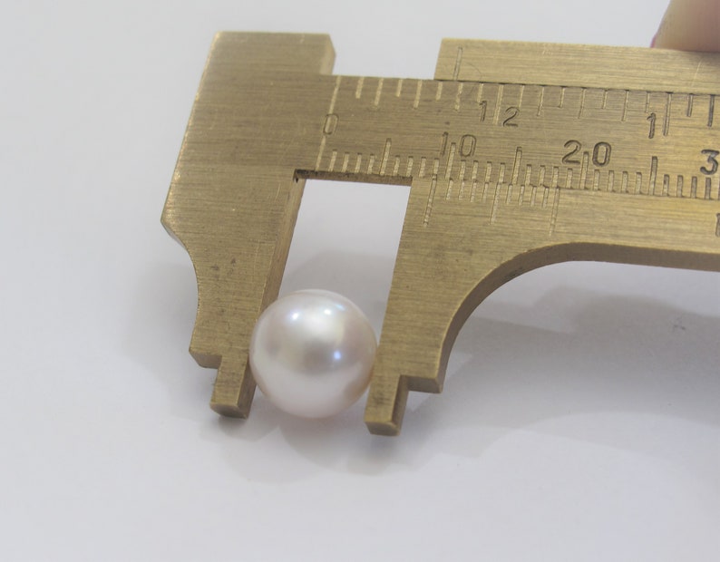 May include: A white pearl is measured with a brass caliper. The pearl is approximately 10 millimeters in diameter.