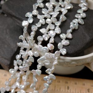 May include: A strand of white, irregularly shaped freshwater pearls. The pearls are strung together, creating a cascading effect. The pearls are displayed on a dark gray stone, with a white teacup and a wooden ruler in the background.