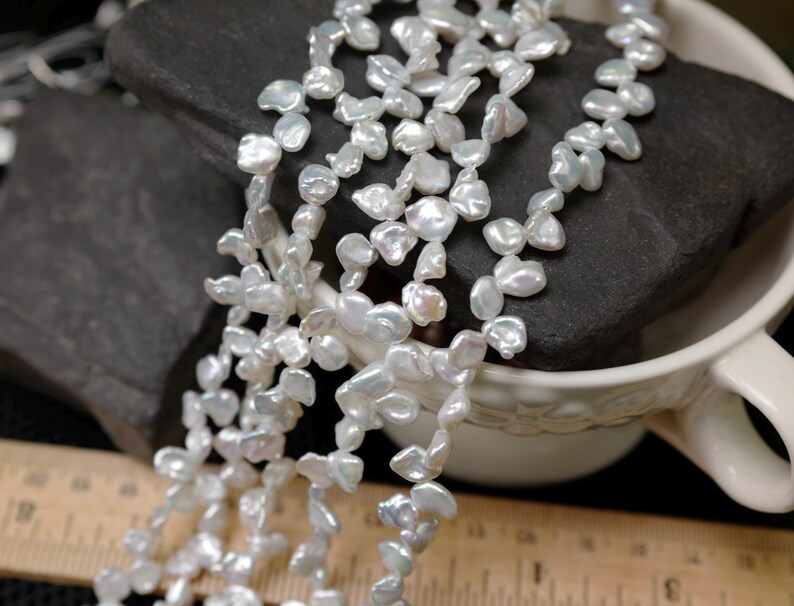 May include: A strand of white, irregularly shaped freshwater pearls. The pearls are strung together, creating a cascading effect. The pearls are displayed on a dark gray stone, with a white teacup and a wooden ruler in the background.