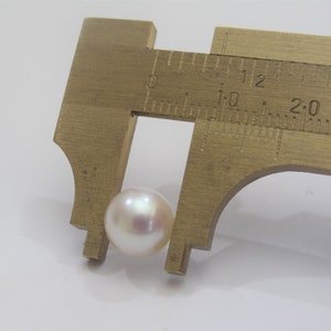 May include: A white pearl measuring approximately 1.2 cm in diameter, being measured with a caliper.