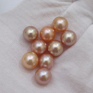 May include: A collection of lustrous, round pearls in varying shades of peach, pink, and gold. The pearls are displayed on a white fabric background, showcasing their smooth surfaces and natural iridescence. The pearls are of different sizes.